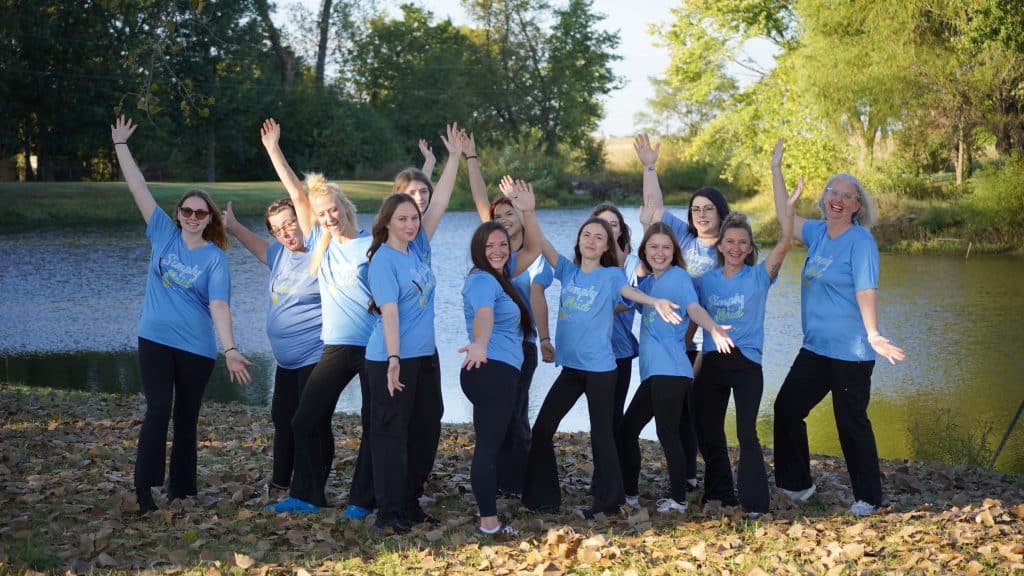 Group of smiling cleaning service team members from Simply Maid, wearing blue shirts, posing joyfully by a pond, celebrating customer service and teamwork.