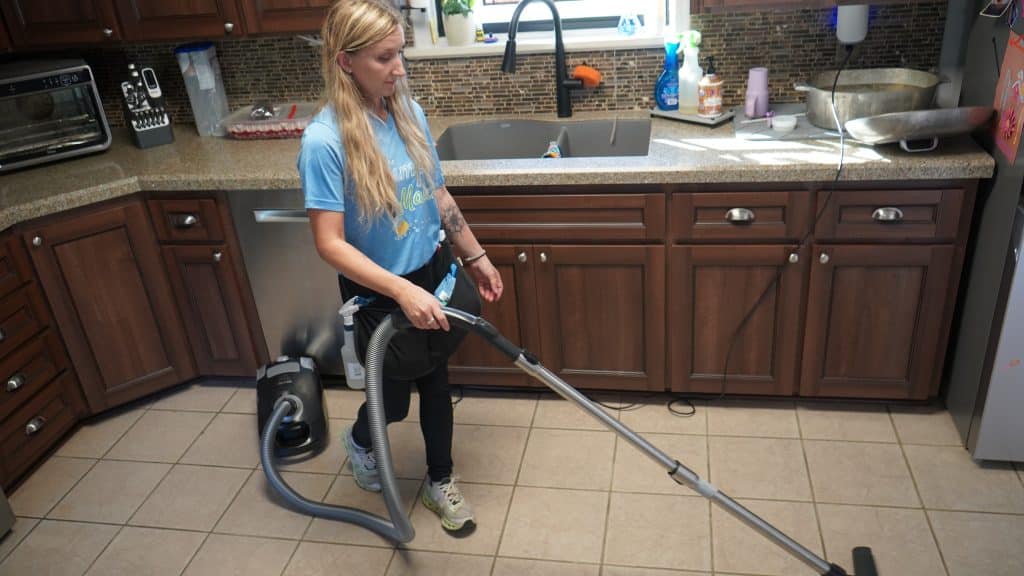 Woman vacuuming kitchen floor in residential setting, showcasing professional cleaning techniques and equipment relevant to cleaning industry education.