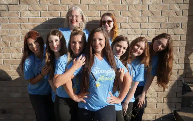 Group of smiling women wearing Simply Maid branded t-shirts, posing together in front of a brick wall, showcasing team spirit and professionalism for a house cleaning service.