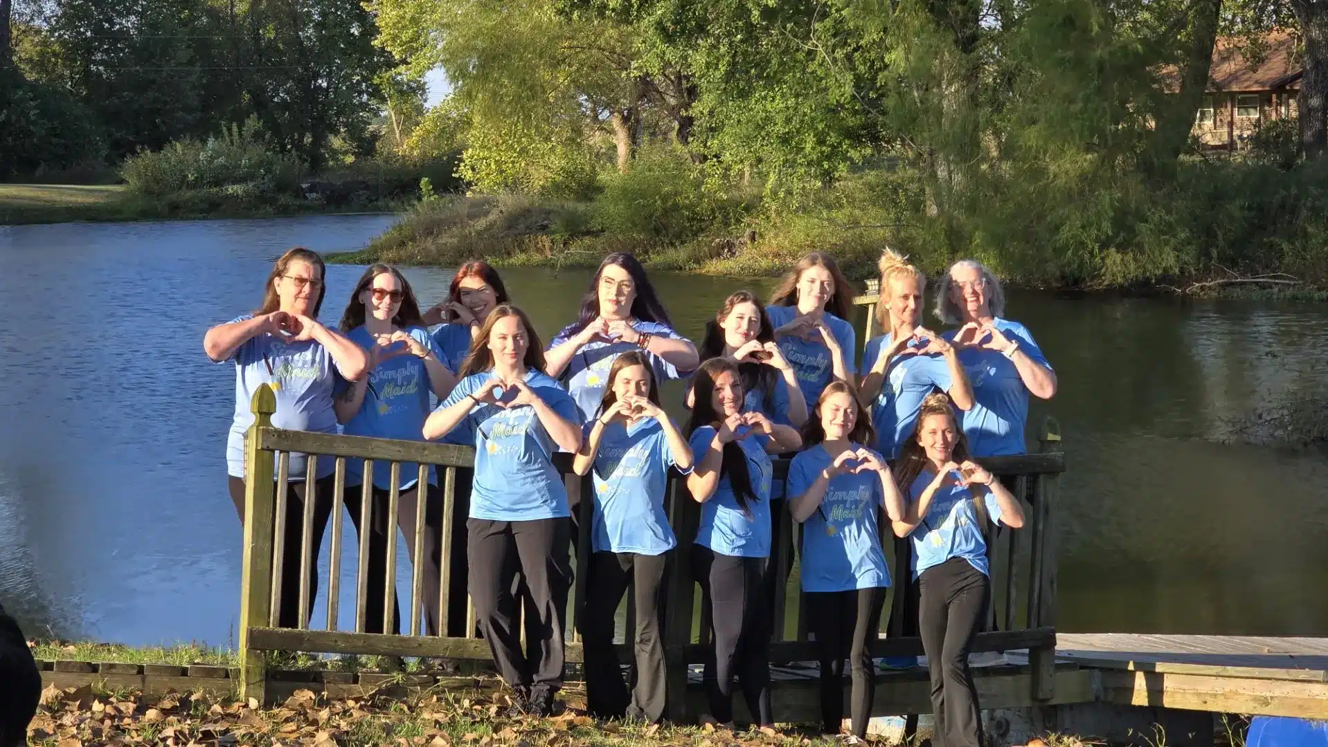 Group of smiling individuals in blue Simply Maid t-shirts forming heart shapes with their hands, standing by a serene water backdrop, promoting community and teamwork in move-out cleaning services.